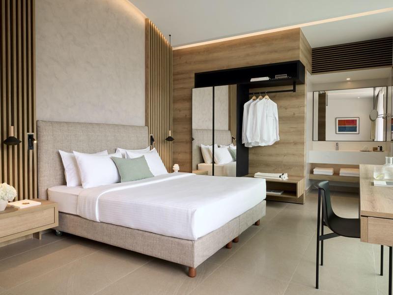 Moderní hotelový pokoj s velkou postelí, pracovním stolem a otevřenou skříní v neutrálních barvách.