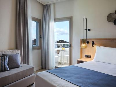 Camera d'hotel luminosa con letto matrimoniale, zona salotto e balcone con vista sul mare.