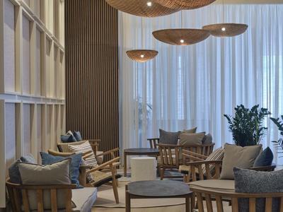 Area salotto moderna con sedie in legno, cuscini grigi e grandi lampade sospese davanti a tende bianche.