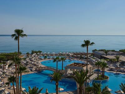 Blick auf einen Hotelpool mit Palmen und Sonnenschirmen am Meer bei klarem Himmel.