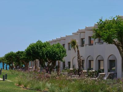 Hotelgebäude mit Terrasse, grünen Bäumen und Meer im Hintergrund bei klarem Himmel.