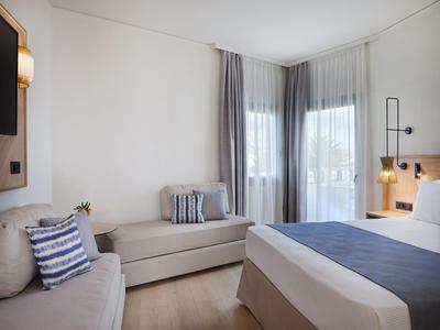 Helles Hotelzimmer mit Bett, zwei Sofas, Kissen und großen Fenstern mit Vorhängen.