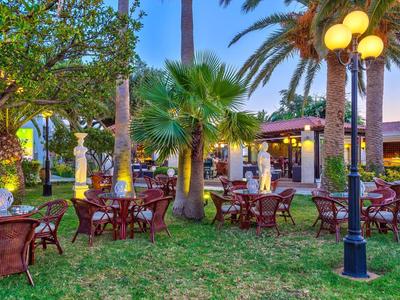 Freiluftterrasse mit Tischen und Stühlen auf Rasen, umgeben von Palmen und Lampen.