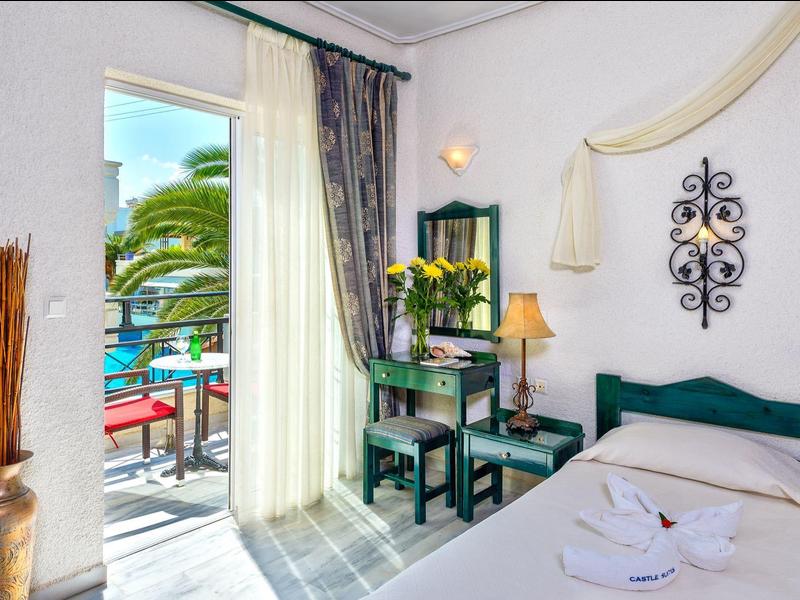 Licht hotelkamer met tweepersoonsbed, bureau en balkon met uitzicht op zee en palmbomen.