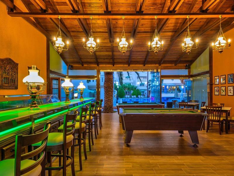 Heldere hotelbar met groene verlichting, barkrukken en pooltafel onder houten balkenplafond.