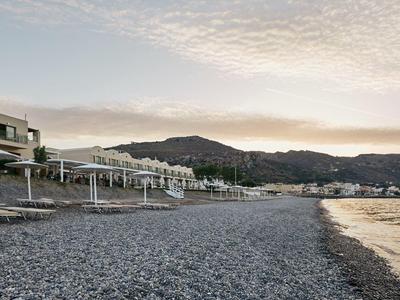 Spiaggia di ciottoli con ombrelloni davanti a edifici moderni e colline sullo sfondo al tramonto
