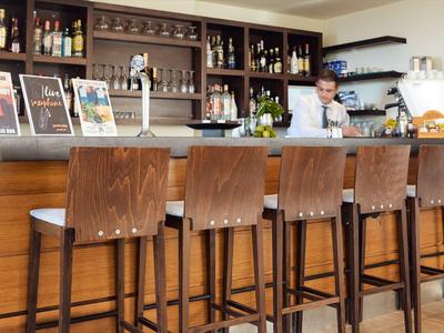 Bar de hotel moderno con mostrador de madera, sillas altas y camarero al fondo.