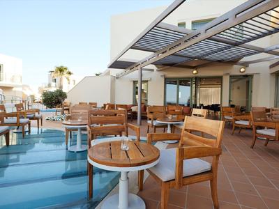 Área exterior de hotel con sillas y mesas de madera junto a una piscina bajo un techo de vidrio.