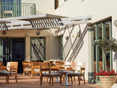 Terraza con mesas y sillas de madera frente a un edificio de hotel con fachada de vidrio y plantas.