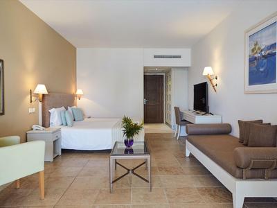 Habitación de hotel luminosa con cama doble, sofá, televisor y mobiliario moderno.
