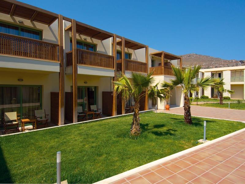 Hotel con camere su due piani, prato e palme sotto un cielo azzurro.