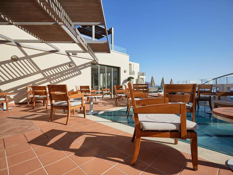 Terrazza soleggiata con sedie e tavoli in legno accanto a una piccola piscina sotto un cielo azzurro.