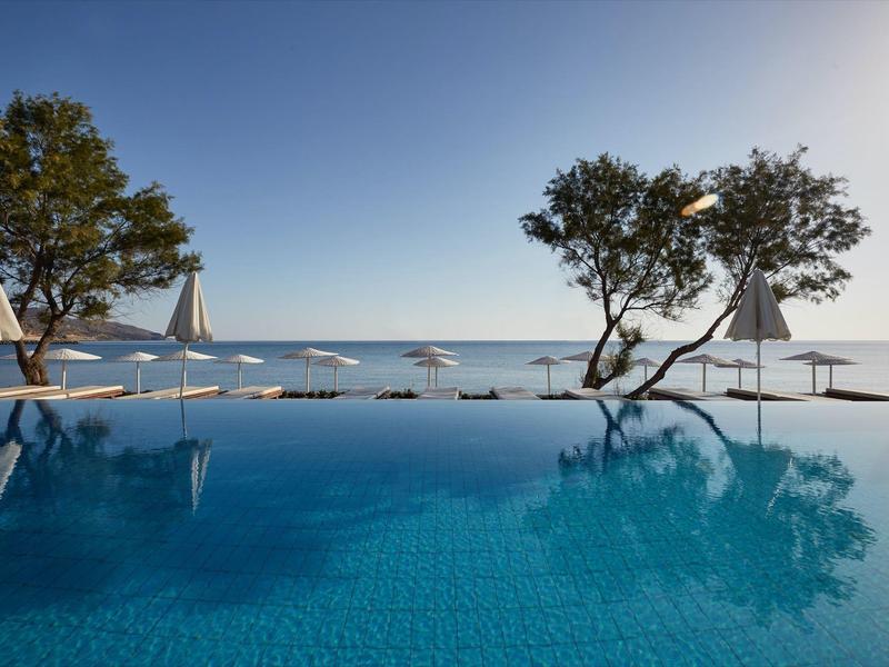 Piscina a sfioro con vista sul mare, circondata da alberi e ombrelloni sotto un cielo limpido.
