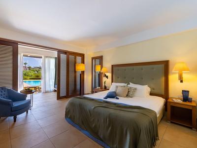 Habitación de hotel luminosa con cama grande, área de estar y vista a una piscina exterior.