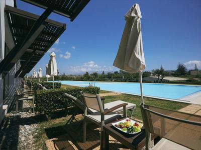Área de piscina con sombrillas, sillas y una mesa con frutas, hotel al fondo.