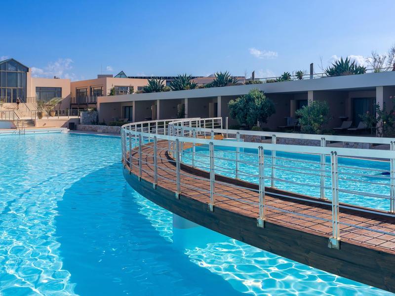 Gran piscina exterior con puente y edificios del hotel alrededor bajo un cielo azul.