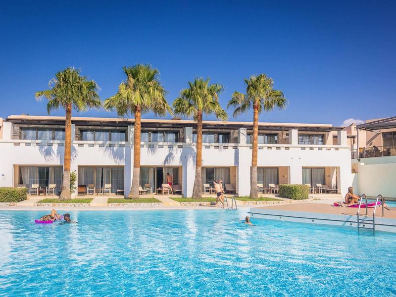 Piscina hotelera moderna con palmeras frente a un edificio de dos pisos bajo cielo azul claro.