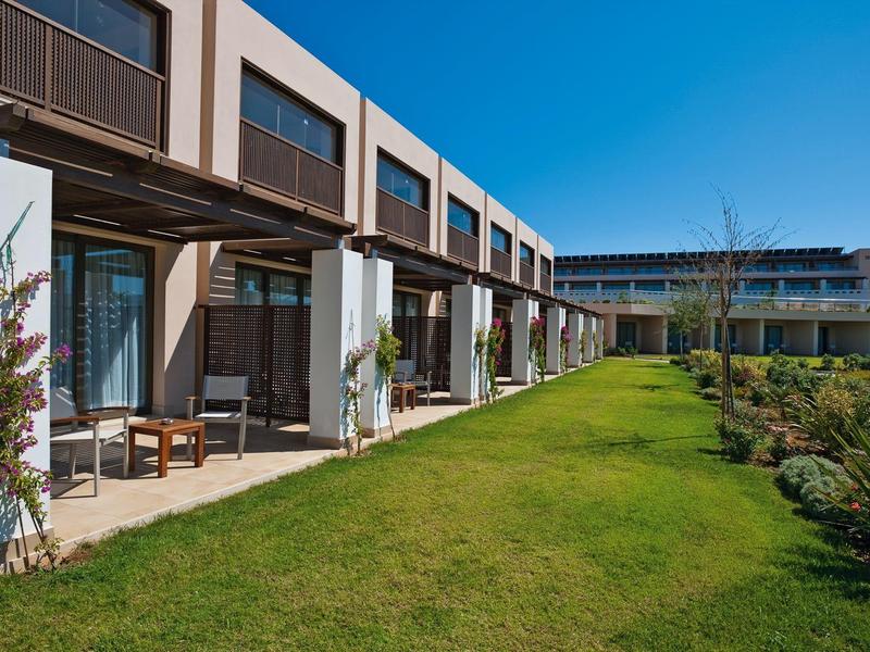 Complejo hotelero moderno con terraza, jardín y sol bajo cielo despejado.