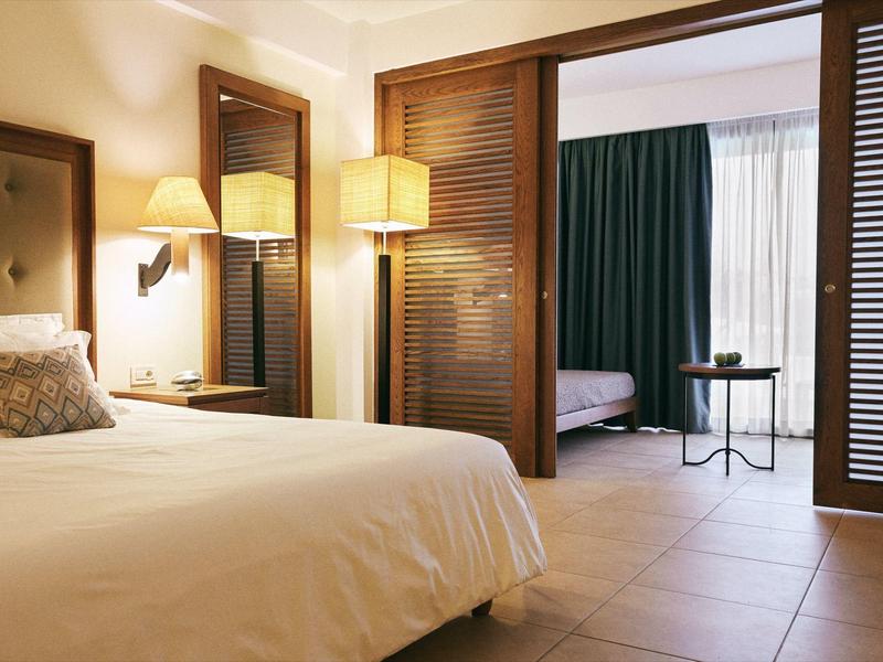 Habitación de hotel luminosa con cama doble, lámparas junto a la cama y puertas de madera abiertas hacia una habitación contigua.