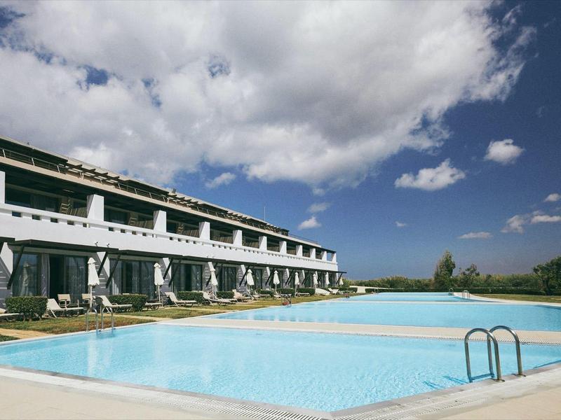 Complejo hotelero moderno con gran piscina al aire libre bajo cielo azul con nubes blancas.
