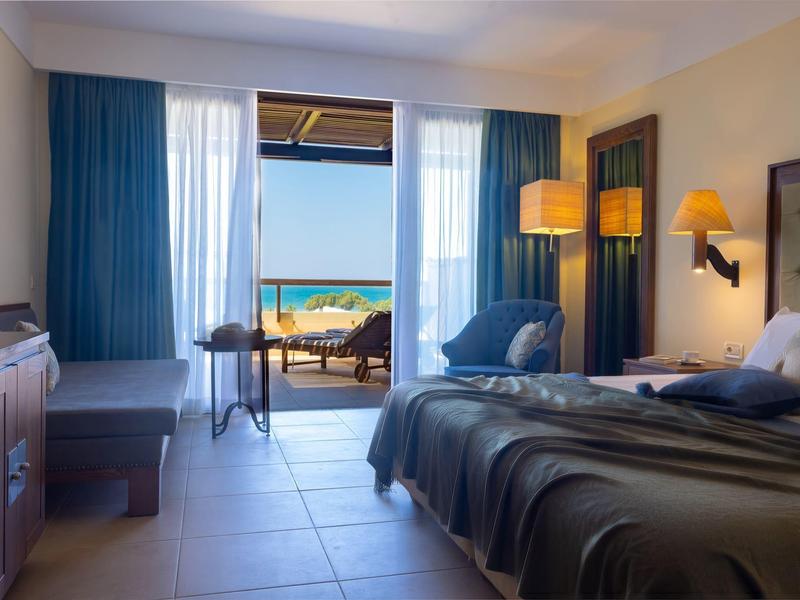 Habitación de hotel luminosa con cama doble, decoración azul y vista a una terraza y el océano.