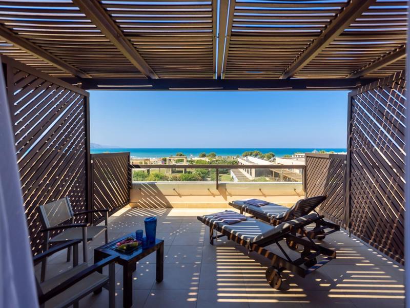 Vista desde la cabaña sombreada en la playa con tumbonas y mar azul al fondo.