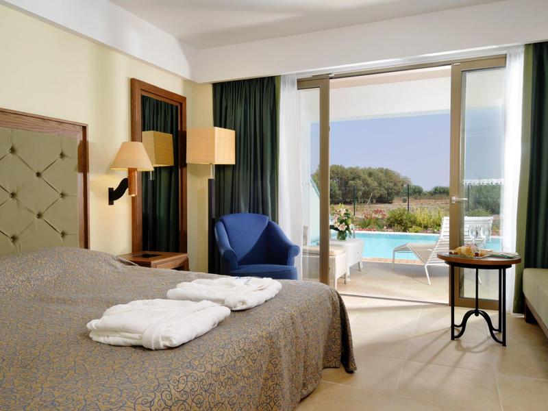 Habitación de hotel con cama doble, dos batas, sillón y vista a la piscina y al mar.