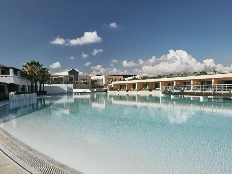 Gran piscina del hotel con agua azul clara rodeada de edificios de dos pisos bajo un cielo nublado.