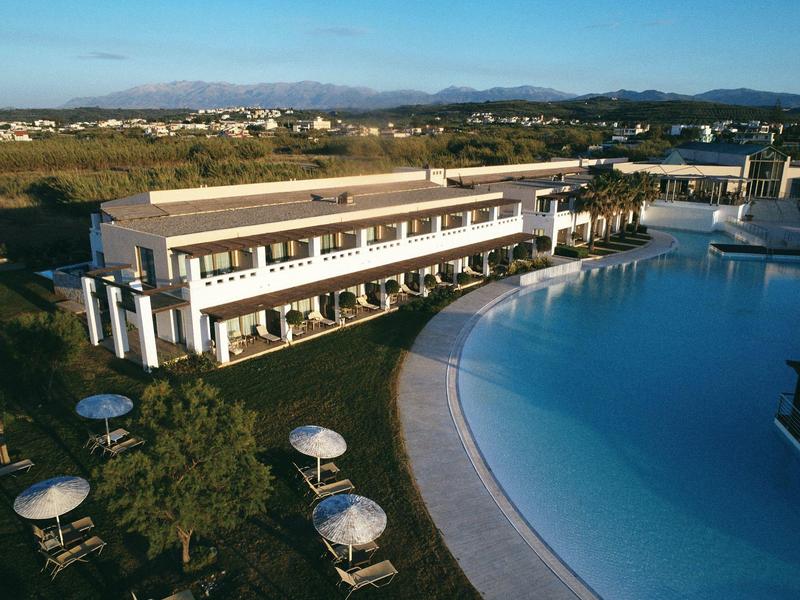 Gran edificio de hotel junto a una gran piscina curva con sombrillas y árboles.