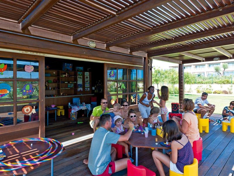 Niños sentados bajo un techo de madera en mesas, haciendo manualidades o jugando juntos.