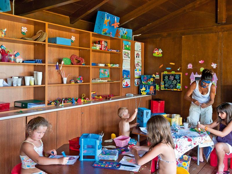 Niños y una cuidadora en una sala de juegos con estanterías llenas de juguetes y libros en un hotel.