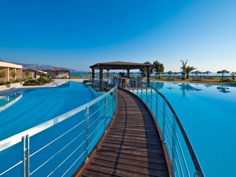 Un amplio muelle conecta dos grandes piscinas azules en un complejo hotelero bajo un cielo claro.
