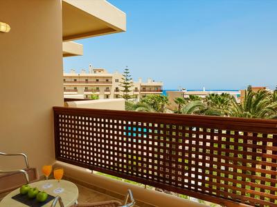Balkon eines Hotels mit Tisch, Stühlen und Blick auf Palmen und Meer bei klarem Himmel.