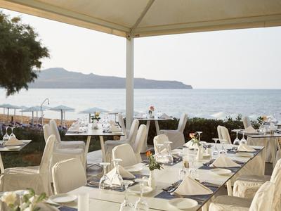 Freiluft-Restaurant mit gedeckten Tischen, weißen Stühlen und Meerblick bei bewölktem Himmel.