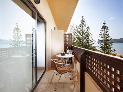 Balkon mit zwei Stühlen, kleinem Tisch, Glasflasche und Blick auf Bäume und Meer im Hintergrund.