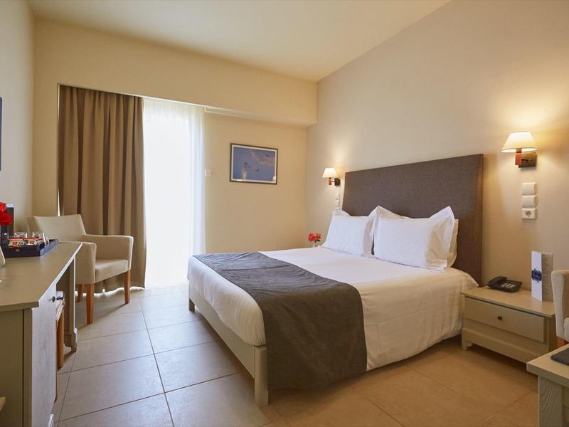 Habitación de hotel luminosa con cama doble, mesitas de noche, escritorio y sillas, colores neutros.