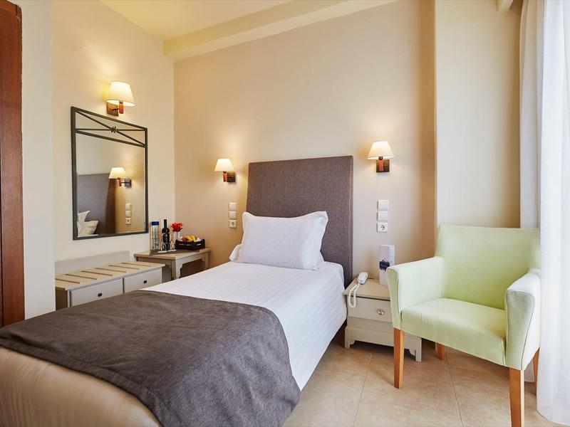 Acogedora habitación individual con cama, espejo, mesa de noche y sillón verde con luz cálida.