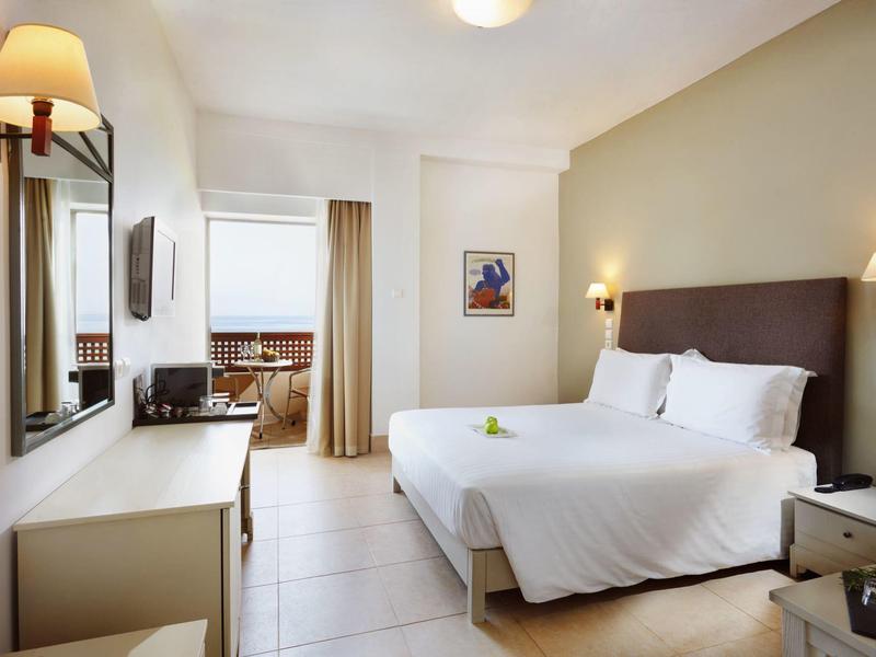 Habitación de hotel luminosa con cama doble, mesitas, escritorio y ventana con cortinas.