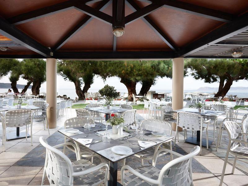 Restaurante en terraza con mesas y sillas blancas, vista al mar y árboles.