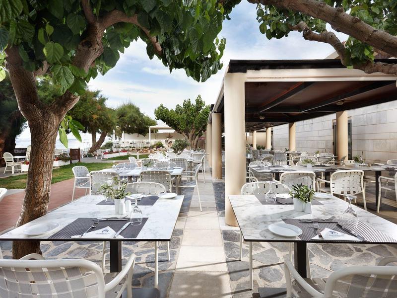 Terraza de restaurante al aire libre con mesas y sillas blancas bajo árboles y un toldo.