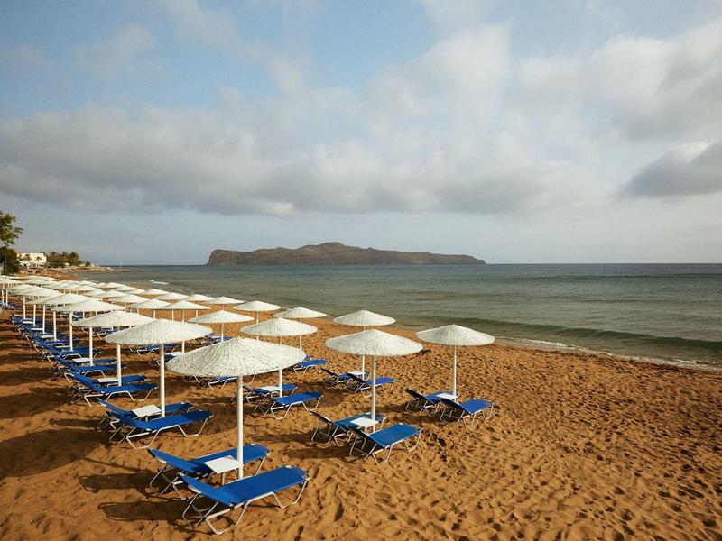 Fila de tumbonas azules vacías y sombrillas blancas en la playa de arena con mar y isla al fondo.