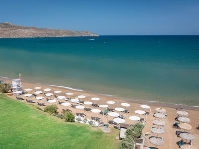 Strand mit weißen Sonnenschirmen, Sand und grünem Gras neben türkisfarbenem, ruhigem Meer.