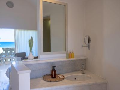 Helles Badezimmer mit weißem Waschbecken, großer Spiegel, Pflanze und Blick auf Meer und Terrasse.