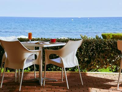 Terrasse mit Tisch, Stühlen und Getränken vor Blick aufs Meer bei sonnigem Wetter.