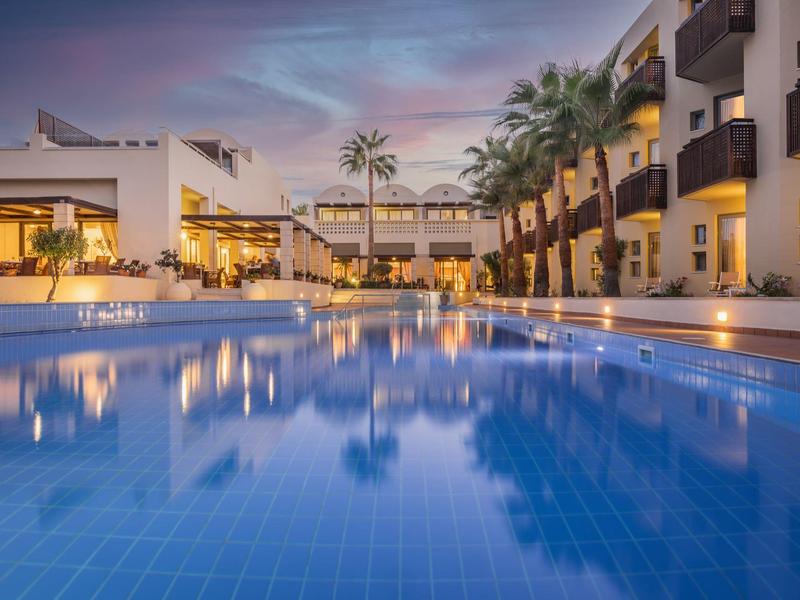 Avondzicht op een resortzwembad met palmbomen en gebouwen weerspiegeld in het water.