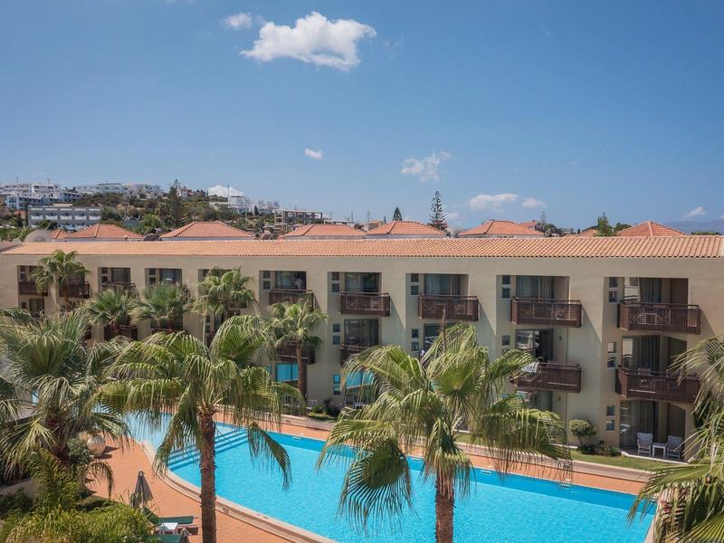 Hotel met balkons en een zwembad omgeven door palmbomen onder een blauwe lucht.