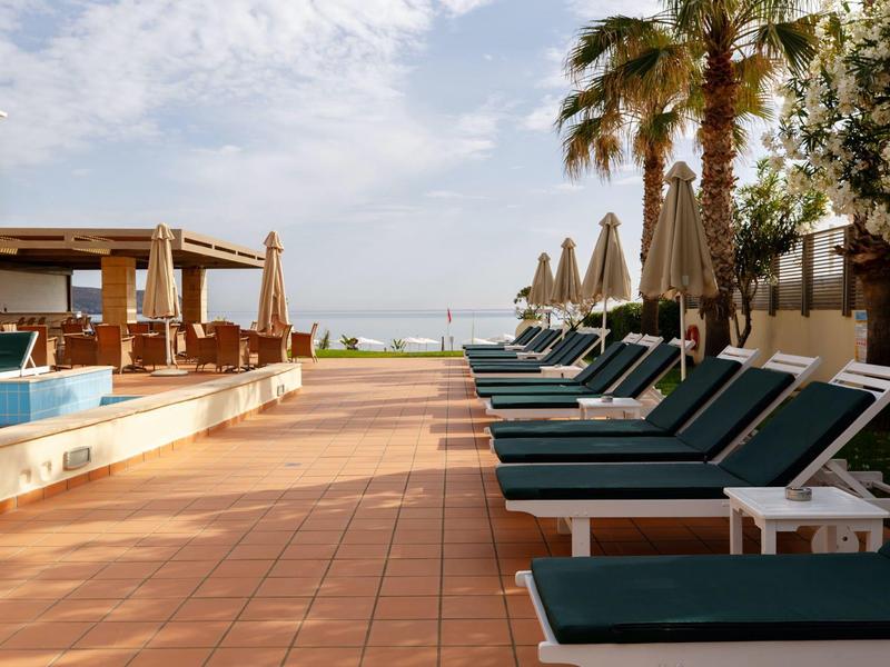 Ligstoelen bij het zwembad, omringd door palmbomen en parasols onder een licht bewolkte hemel.