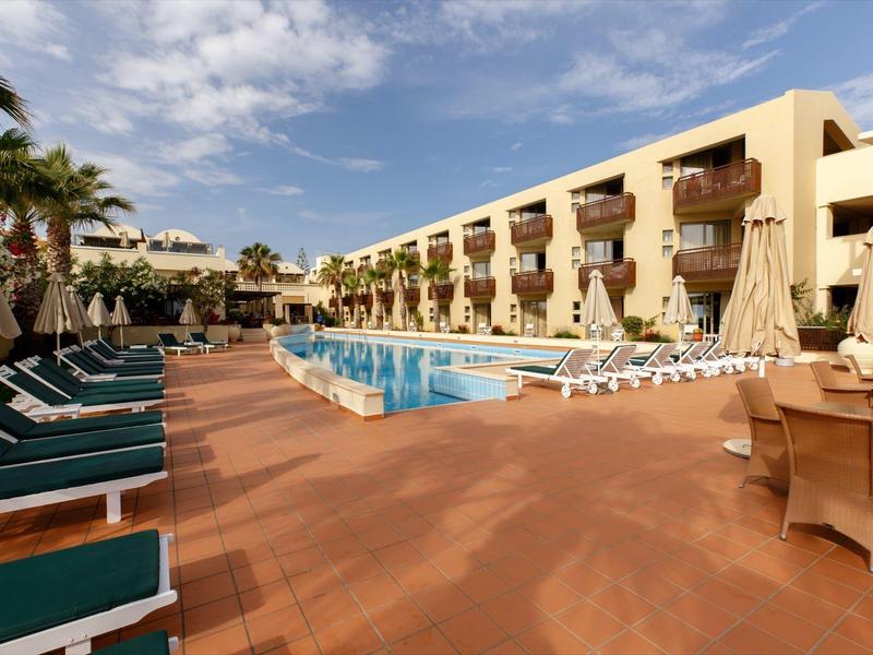 Hotelzwembad met ligbedden, parasols en balkons onder een blauwe lucht.