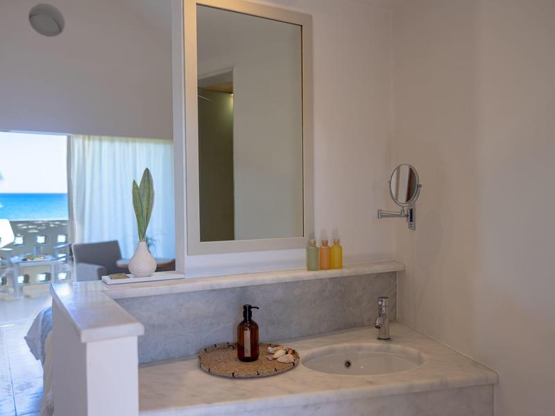 Lichte badkamer met grote spiegel, witte muren en uitzicht op zee buiten.