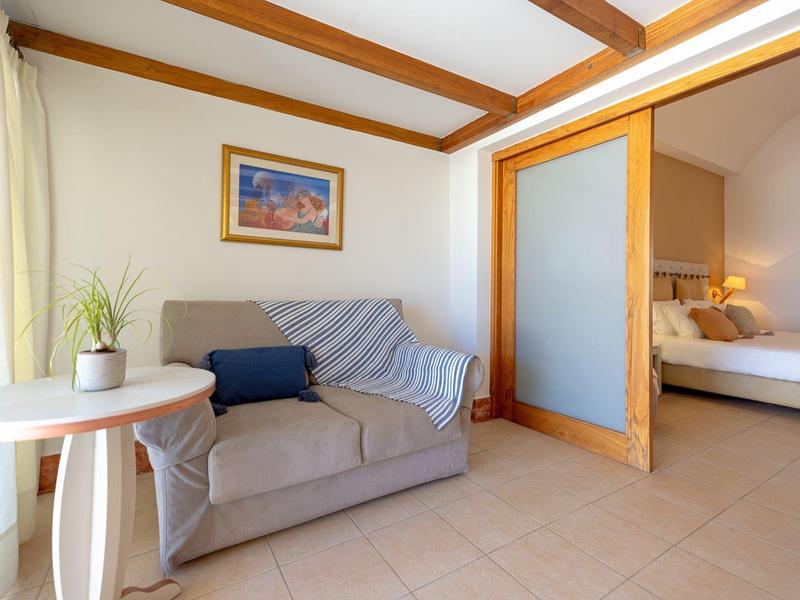 Gezellige zithoek met grijze bank, blauw kussen, witte ronde tafel en slaapkamer achter schuifdeur.